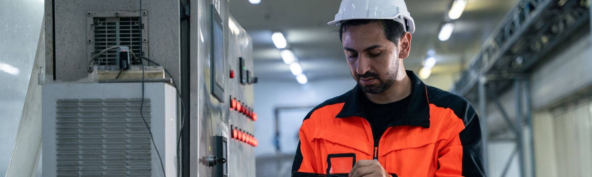 Engineer in high visibility workwear using a tablet while checking controls inside a manufacturing facility.