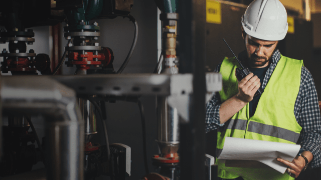 Engineer in high-visibility vest and hard hat using a radio while reviewing technical plans in an industrial facility.