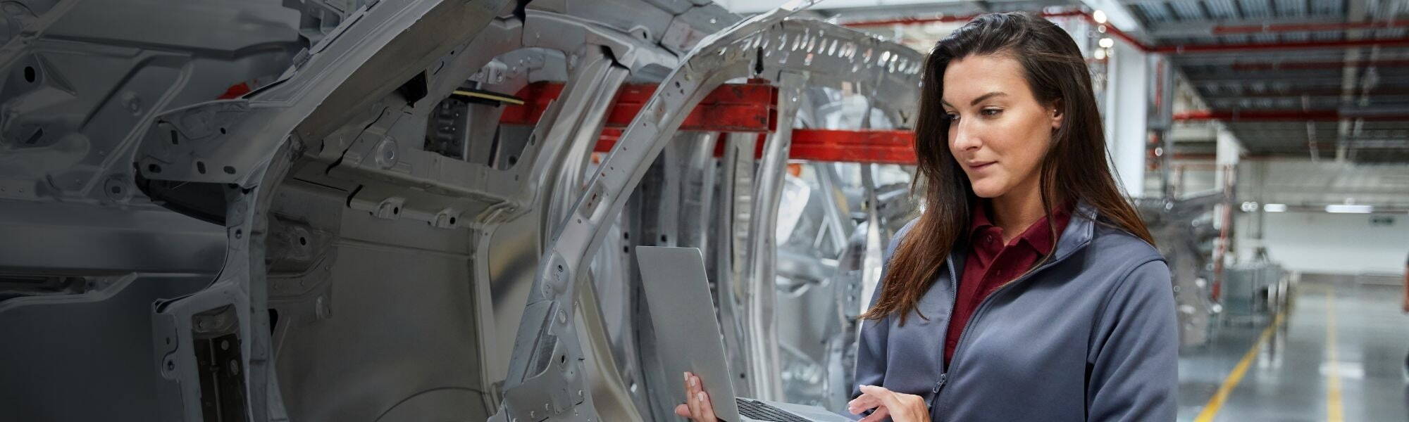 Female automotive engineer inspecting car chassis structure with a laptop in a modern vehicle manufacturing plant.