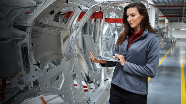 Female automotive engineer inspecting car chassis structure with a laptop in a modern vehicle manufacturing plant.
